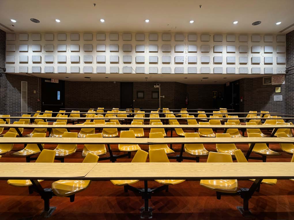Photo of an empty tiered seating lecture hall with yellow seats and red carpet.