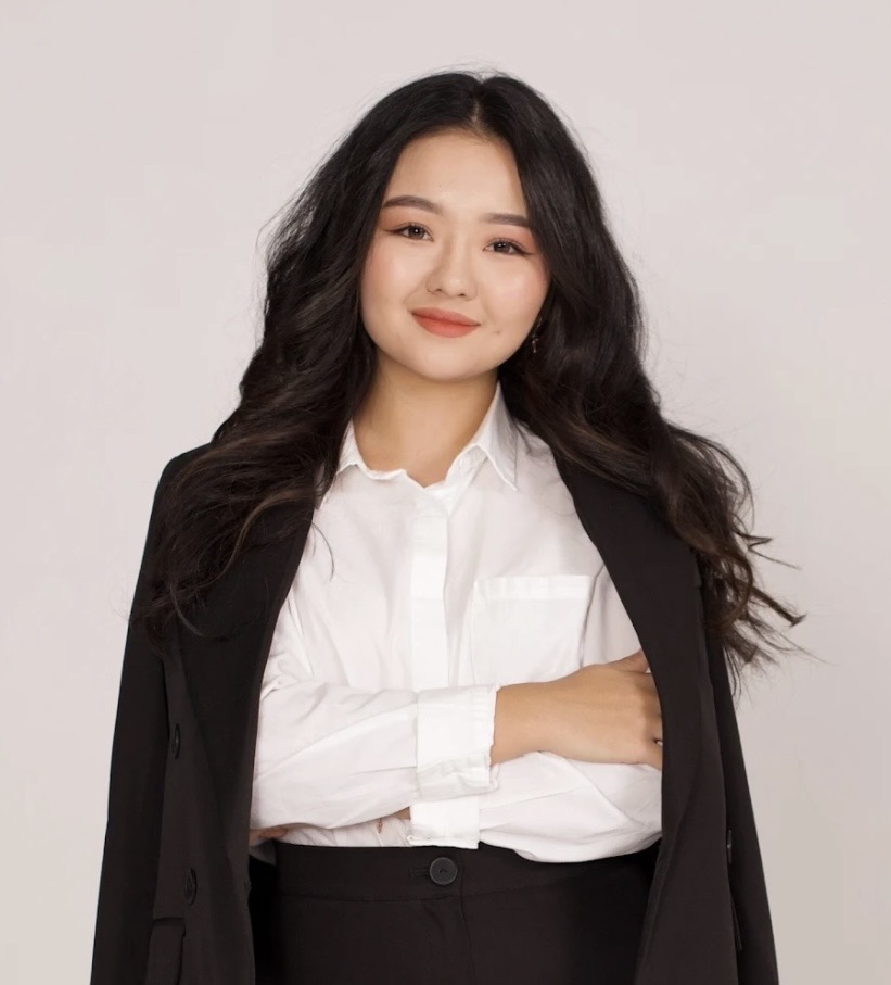 Ainel Demegenova Smiling young woman with long, wavy, dark hair, a black coat over her shoulders, wearing a white blouse and black bottoms with her arms crossed