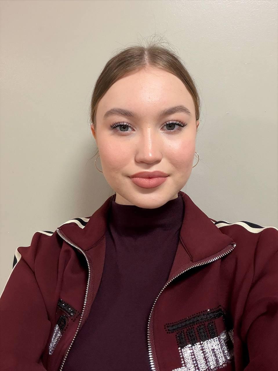 Young woman with tied back blonde hair wearing a burgundy high neck top and a track suit jacket against a beige wall