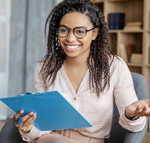 A smiling young woman with long, curly, black hair and glasses, wearing a beige blouse, sits and genstures broadly while holding a blue clipboard and a pen.