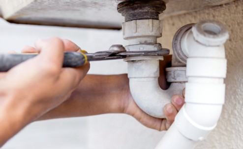 Hands holding a wrench, working on a pipe under a sink.