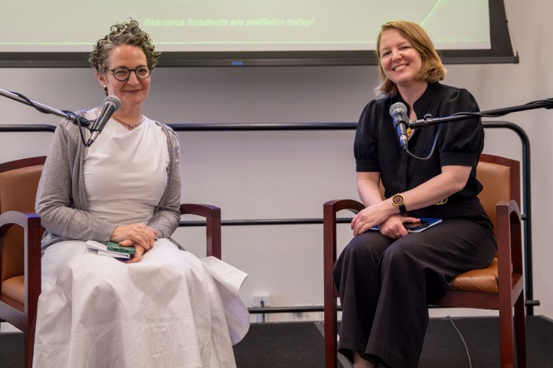 Two smiling women, one in a white dress and a grey cardigan with short, wavy hair and dark-rimmed glasses, the other in a black blouse and trousers with shoulder-length red hair, sit in orange and brown chars with a presentation projected behind them and microphones in front of them.