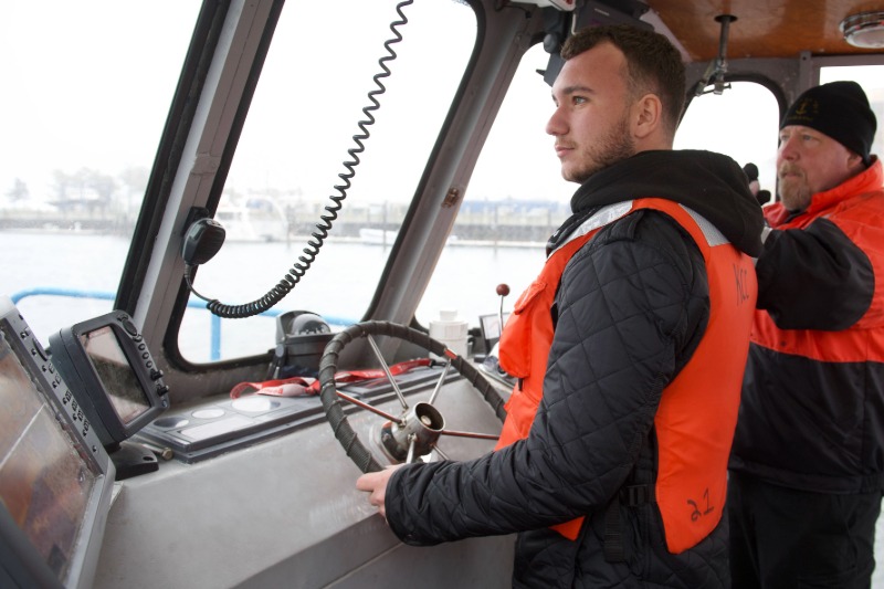 Two men in a boat wearing high-visibility black and orange clothing, one steering, the other pointing something out.